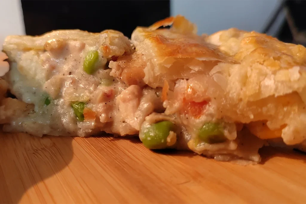 A slice of flaky chicken pot pie with puff pastry on a wooden food board